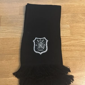 ”Svart AIK halsduk med gamla skölden” - Svart halsduk från AIK med klubbens klassiska märke broderat framtill. Halsduken har långa tofsar i ändarna och är perfekt för att visa ditt stöd på läktaren. Officiell merchandise från AIKshop.se. Pris kan diskuteras 