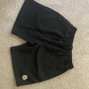 Svarta shorts från Moncler med snörning - Svarta Moncler shorts med elastisk midja och snörning med metalländar där Moncler är ingraverat. Klassisk logga på vänster ben. Sköna och stilrena, perfekta för sommaren. Tillverkade i mjuk bomull för maximal komfort och en loose passform.
