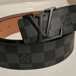 Svart Louis Vuitton bälte - Snyggt svart bälte från Louis Vuitton med ett klassiskt rutigt mönster. Bältet har en stilren svart spänne med LV-logotypen. Perfekt för att ge en lyxig touch till din outfit.
