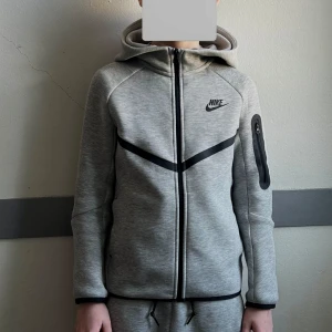 Grå tech fleece hoodie från Nike - Säljer en nästan helt oanvänd grå Nike tech fleece hoodie från Nike med dragkedja och huva. Tröjan har svarta detaljer och en ficka med dragkedja på ärmen. Köpt på Kidsbrandstore. Den är i barnstorlek M (137-147cm). Priset är inte hugget i sten för rätt köpare.