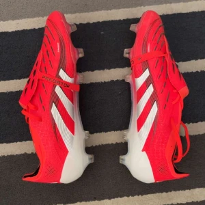 Röda Adidas Predator fotbollsskor - Säljer ett par röda Adidas Predator fotbollsskor med vita och svarta detaljer. Skorna har snörning och är designade för optimal prestanda på planen. Perfekta för fotbollsspelare som vill ha både stil och funktion.