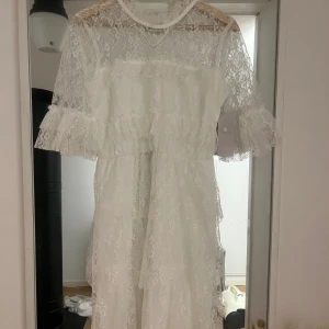 Vit spetsklänning - Elegant vit spetsklänning med trekvartsärm och volangdetaljer. Perfekt för sommarens festligheter. Klänningen är endast testad. STL 34