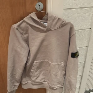 Beige hoodie från Stone Island - Säljer en snygg beige hoodie från Stone Island med deras ikoniska märke på ärmen. Tröjan har en stor ficka framtill och en bekväm huva. Perfekt för en avslappnad stil. Skick 10/10 och Storlek 12år