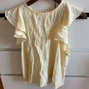 Gul topp med volangärmar från Zara - Säljer en söt gul topp från Zara med volangärmar och en öppen ryggdetalj. Perfekt för varma dagar och ger en romantisk touch till din outfit. Tillverkad i ett lätt och luftigt material.
