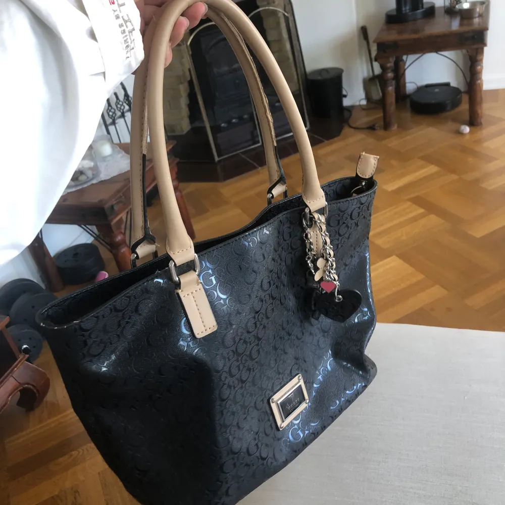 Snygg svart handväska från Guess med beige handtag och ett stilrent mönster av logotypen. Väskan har en metallplatta med märkesnamnet och en charmig nyckelring med hjärta. Insidan är rymlig med ett matchande mönster.. Laukut & Käsilaukut.