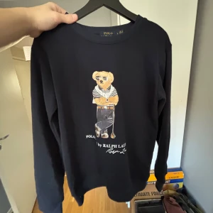 Svart tröja med björntryck från Ralph Lauren - Säljer en svart långärmad tröja från Ralph Lauren med ett fint björntryck på framsidan. Perfekt för dig som gillar stilrena och unika plagg. Tröjan har en klassisk passform och är gjord i ett mjukt material. 