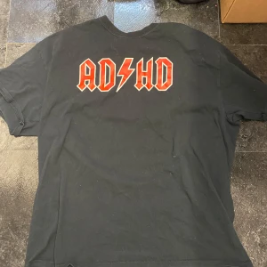 Svart t-shirt med ADHD-tryck - Cool svart t-shirt med ett iögonfallande ADHD-tryck i rött och vitt. Perfekt för dig som vill sticka ut med en unik stil. T-shirten har korta ärmar och en normal passform.