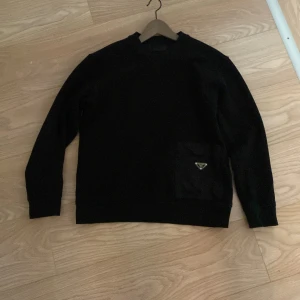 Svart tröja från Prada - Snygg svart tröja från Prada med en praktisk ficka på framsidan. Tröjan har långa ärmar och en klassisk rund halsringning. Perfekt för en stilren look.