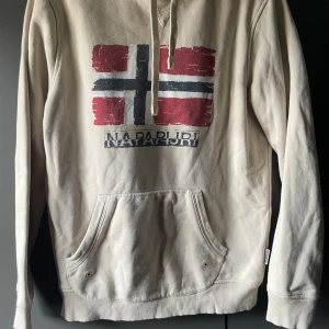 Beige hoodie från Napapijri - Säljer en beige hoodie från Napapijri med en stor norsk flagga tryckt på framsidan. Tröjan har en känguruficka och justerbar huva med snören. Perfekt för en avslappnad stil.