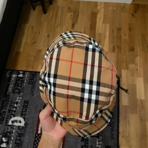 Rutig hatt från Burberry - Snygg rutig hatt från Burberry i klassiskt beige, svart och rött mönster. Perfekt för att ge din outfit en stilfull touch. Hatten har en bred kant och är tillverkad i ett lätt material.