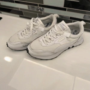 Vita sneakers från Puma - Snygga vita sneakers från Puma med klassisk design och svarta sulor. Skorna har snörning och en bekväm passform, perfekt för vardagsbruk. Puma-loggan syns tydligt på sidan och hälen.