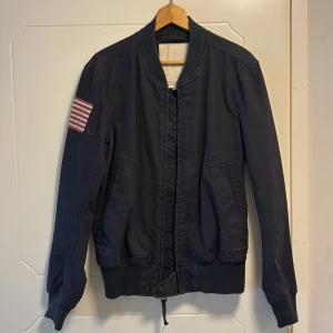 Mörkblå bomberjacka från Ralph Lauren - Säljer en snygg mörkblå bomberjacka från Ralph Lauren med en cool amerikansk flagga på ärmen. Jackan har en dragkedja framtill och ribbade muddar vid ärmslut och nederkant. Perfekt för en stilren look!