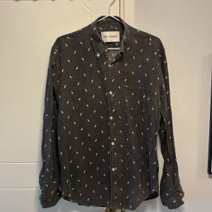 Grå skjorta från Our Legacy - Snygg grå skjorta från Our Legacy med ett diskret mönster. Skjortan har långa ärmar och knappar framtill. Perfekt för en stilren look.