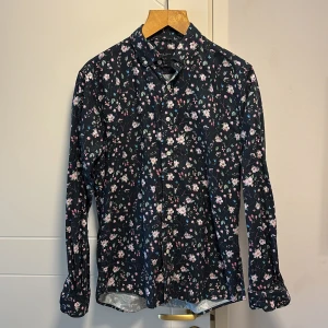 Blommig skjorta från Grand Frank - Snygg blommig skjorta från Grand Frank i en mörk färg med ljusa blommor. Skjortan har en slim fit passform och långa ärmar, perfekt för en stilren look. Knappar framtill ger en klassisk touch.