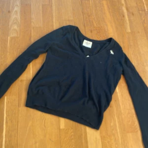 Svart tröja från Abercrombie & Fitch - Säljer en stilren svart tröja från Abercrombie & Fitch. Tröjan har en v-ringad design, perfekt för en avslappnad look. Den är tillverkad i ett mjukt material som ger en bekväm passform. En liten logga syns på bröstet.