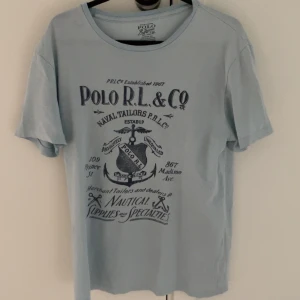 Ljusblå t-shirt från Polo R.L. & Co - Säljer en ljusblå t-shirt från Polo R.L. & Co med ett stort tryck på framsidan. T-shirten har en rund hals och korta ärmar. Perfekt för en avslappnad stil.