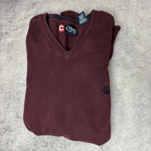 Vinröd ärmlös tröja från Chaps - Säljer en stilren vinröd ärmlös tröja från Chaps i storlek M. Tröjan har en v-ringad design och är tillverkad i 100% bomull. Perfekt för lager-på-lager-stil eller som ett elegant inslag i garderoben.