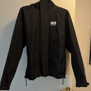 Svart vindjacka från Helly Hansen - Säljer en svart vindjacka från Helly Hansen med huva och dragkedja. Jackan har långa ärmar och justerbar nederkant. Perfekt för utomhusaktiviteter och skyddar mot vind och regn. 4 fickor.