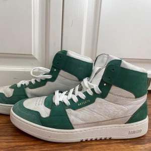 Vita och gröna sneakers från Axel Arigato - Snygga vita och gröna sneakers från Axel Arigato med hög design och snörning. Skorna har en stilren look med märkets logga på sidan och sulan. Perfekta för en trendig stil. Ingen box.