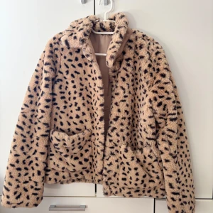 Beige pälsjacka från H&M Divided - Säljer en snygg beige pälsjacka från H&M Divided med svart leopardmönster. Jackan har långa ärmar och knappar framtill.