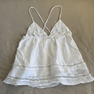 Vit ärmlös topp från Gina Tricot - Säljer en söt vit ärmlös topp från Gina Tricot med tunna axelband och dekorativa spetsdetaljer. Perfekt för varma sommardagar. Toppen har en luftig passform och korsade band i ryggen.