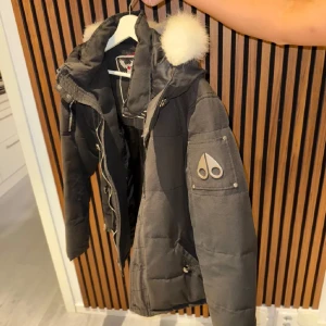 Svart dunjacka med pälsdetaljer - Snygg svart dunjacka från Moose Knuckles med vita pälsdetaljer på huvan. Jackan har en dragkedja och knappar framtill samt en logotyp på ärmen. Perfekt för kyliga dagar.