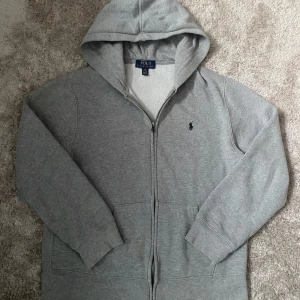 Grå hoodie från Polo Ralph Lauren - Säljer en stilren grå hoodie från Polo Ralph Lauren med dragkedja och broderad logga på bröstet. Tröjan är i superfint skick utan defekter. Hör av er vid funderingar och frågor!