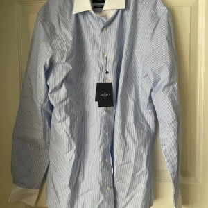 Blå och vit randig skjorta från Hackett - Snygg blå och vit randig skjorta från Hackett i 100% ekologisk bomull. Skjortan har en klassisk krage och långa ärmar med vita manschetter. Perfekt för en stilren look.