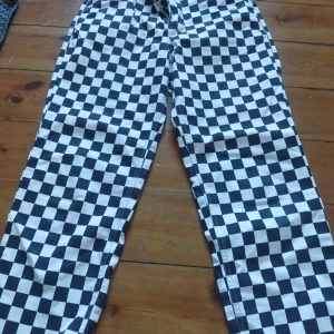 Svartvita rutiga byxor från Vans - Snygga svartvita rutiga byxor från Vans i storlek 27. Byxorna har en klassisk checkerboard-design och är perfekta för en cool och avslappnad stil. De har knappar och dragkedja framtill för enkel på- och avtagning.