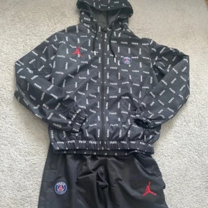 Svart vindjacka med Paris-mönster - Snygg svart vindjacka och byxor från Jordan med Paris-mönster och PSG-logga. Jackan har en dragkedja framtill och en justerbar huva. Perfekt för sportiga tillfällen eller som en stilren vardagsjacka. Använd bara ett fåtal gånger.