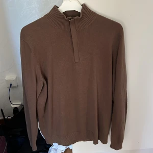 Brun tröja med dragkedja från Boss - Säljer en stilren brun tröja från Boss med dragkedja och ribbad krage. Tröjan har långa ärmar och förstärkta armbågar i matchande färg. Perfekt för en avslappnad men elegant look.