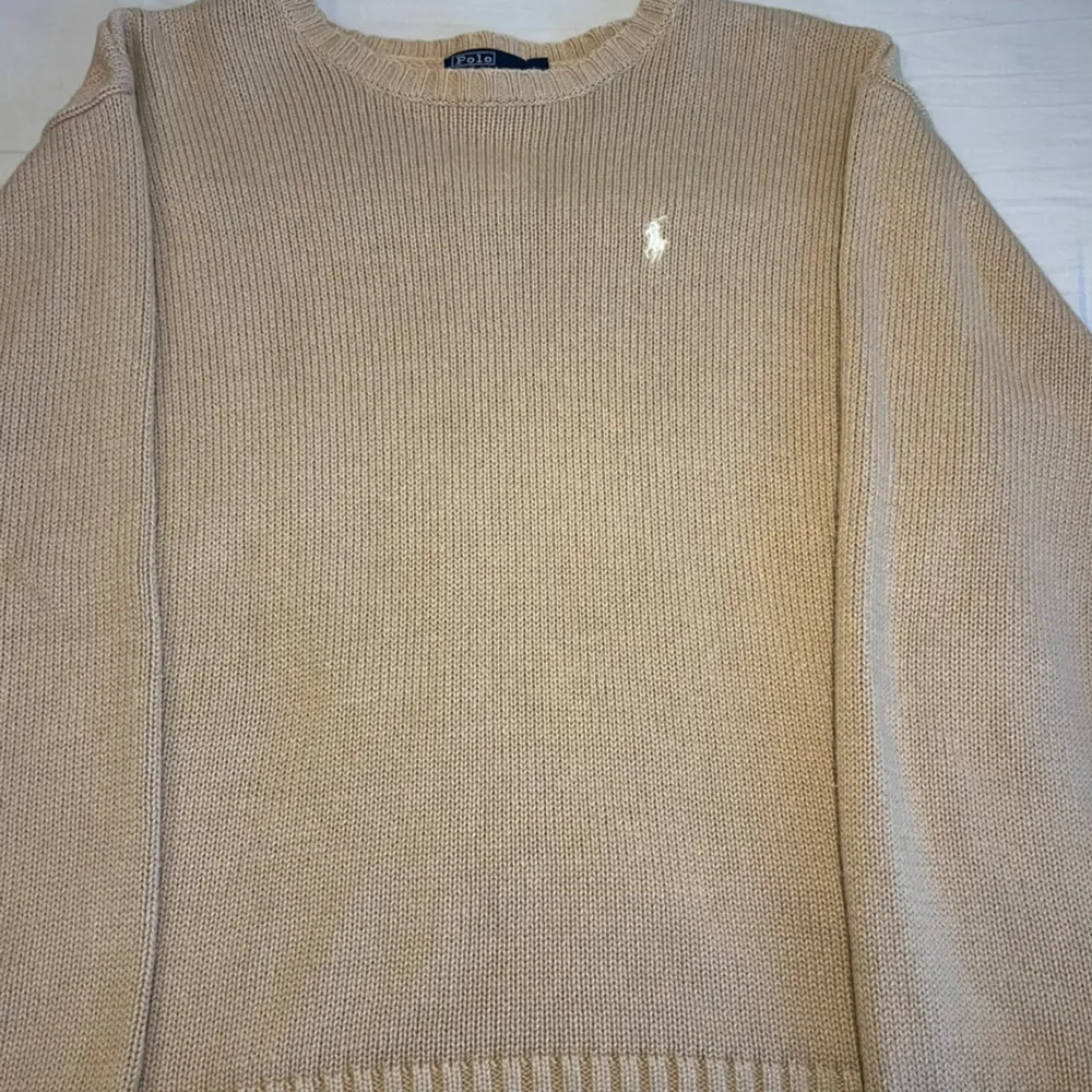 Säljer en stilren beige stickad tröja från Ralph Lauren med det klassiska broderade logotypen på bröstet. Tröjan har långa ärmar och en rund halsringning. Perfekt för en avslappnad och elegant look.. Neuletakit & Villapaidat.