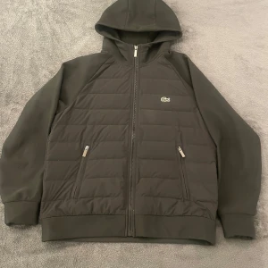 Svart cardigan jacka från Lacoste - Säljer en stilren svart quiltad jacka från Lacoste med huva och dragkedja. Jackan har två praktiska fickor med dragkedjor och det ikoniska Lacoste-märket på bröstet. Perfekt för kyligare dagar!🤩🙌
