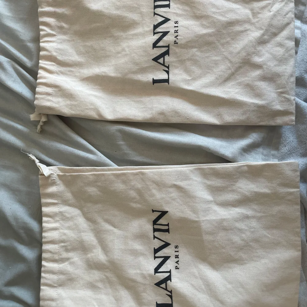 Två eleganta skopåsar från Lanvin i beige med svart text. Perfekta för att skydda dina skor vid förvaring eller resor. Påsarna har en praktisk dragsko för enkel stängning.. Kengät.