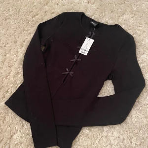 Svart topp med rosetter från Gina Tricot - Snygg svart topp från Gina Tricot med dekorativa rosetter framtill. Perfekt för en stilren look. Toppen har långa ärmar och en elegant design som passar till många tillfällen. Helt ny och aldrig använt. Fick i julklapp men har andra finare tröjor så därför säljer jag denna. Skriv för fler bilder. Pris kan diskuteras 