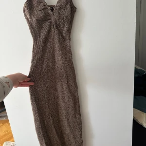 Brun stickad klänning från ADOORE - Säljer en snygg brun stickad klänning från ADOORE med smala axelband och v-ringning. Klänningen har en åtsittande passform och är perfekt för en stilren look. Den är ribbad och har en knytning i ryggen för extra detalj.