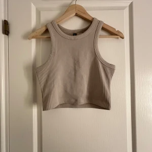 Beige ribbad linnetopp från H&M Divided - Säljer en stilren beige ribbad linnetopp från H&M Divided. Toppen är ärmlös och har en croppad passform, perfekt för varma dagar. Den är enkel att matcha med olika outfits och ger en avslappnad look.