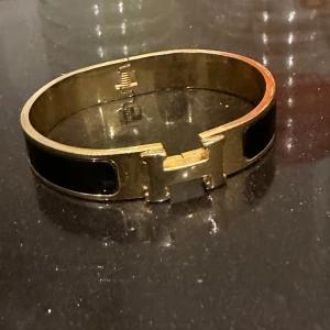 H Guldarmband med svart emalj - Elegant guldarmband med svart emaljdetalj och ett stilrent H-format spänne. Perfekt för att ge en lyxig touch till din outfit.