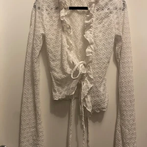 Vit spetsblus från Gina Tricot - Säljer en elegant vit spetsblus från Gina Tricot. Blusen har långa ärmar och fina volangdetaljer framtill. Den är knytbar i midjan för en snygg passform. Perfekt för att ge en romantisk touch till din outfit. Den är i helt ny skick 