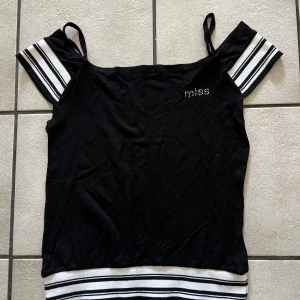 Svart topp med randiga detaljer - Snygg svart topp med smala axelband och randiga detaljer i vitt och svart på ärmarna och nederkanten. Ordet 'miss' är dekorerat med små stenar på bröstet. Perfekt för en trendig look!