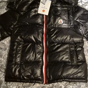 Svart dunjacka från Moncler - Säljer en snygg svart dunjacka från Moncler med glansig finish och ikonisk logga på bröstet. Jackan har en dragkedja i rött, vitt och blått samt praktiska fickor. Perfekt för kyliga dagar! Helt ny men den är lite liten på mig så jag säljer den priset kan sänkas. men har ni några frågor kan skriva.