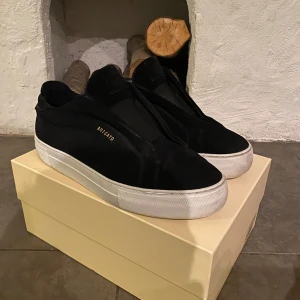 Svarta skor från Axel Arigato - Snygga svarta sneakers från Axel Arigato med en stilren design. Skorna har en vit sula som ger en fräsch kontrast och är tillverkade i mocha och skinn. Perfekta för en trendig look. Skorna är i bra skick. Skriv vid funderingar eller fler bilder!