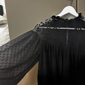 Svart blus från Zara - Elegant svart blus från Zara med transparenta detaljer och diskret mönster. Blusen har långa ärmar och en hög hals med volangkant. Perfekt för en stilren look.