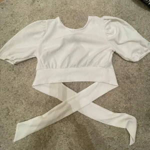Vit topp från H&M Divided - Säljer en vit topp från H&M Divided med korta puffärmar och knytband i midjan. Toppen har en rund halsringning och är tillverkad i ett lätt och bekvämt material. Perfekt för en stilren look!