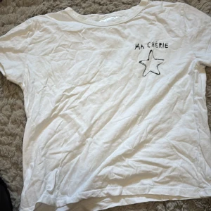 Vit t-shirt med tryck från Gina Tricot - Säljer en vit t-shirt från Gina Tricot med texten 'MA CHÉRIE' och en stjärna tryckt på både fram- och baksidan. Perfekt för en avslappnad stil. 💫