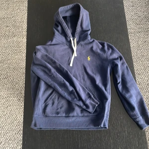 Blå hoodie från Ralph Lauren - Säljer en snygg blå hoodie från Ralph Lauren med en klassisk känguruficka och vit dragsko i huvan. Den har en liten gul logga på bröstet. Perfekt för en avslappnad stil.