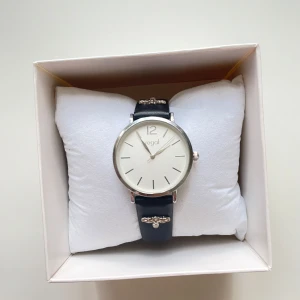 Elegant klocka från Regal - Stilren klocka från Regal med en vit urtavla och silverfärgade detaljer. Klockan har ett svart läderarmband med dekorativa detaljer i silver. Perfekt för en elegant look. Batteriet behöver bytas ut.