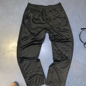 Svarta mjukisbyxor från Adidas - Snygga svarta mjukisbyxor från Adidas med diskret mönster och elastisk midja. Byxorna har en avslappnad passform och är perfekta för en bekväm stil. Adidas-loggan är broderad på sidan.