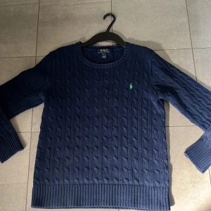 Blå stickad tröja från Ralph Lauren - Säljer en snygg blå stickad tröja från Ralph Lauren med kabelstickat mönster. Tröjan har långa ärmar och en liten grön logga på bröstet. Perfekt för kyligare dagar!
