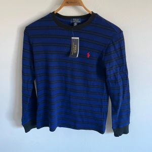 Blå och svart randig tröja från Polo Ralph Lauren - Säljer en snygg blå och svart randig tröja från Polo Ralph Lauren. Tröjan har långa ärmar och en klassisk rund halsringning. Den är dekorerad med det ikoniska röda Polo-logotypen på bröstet. Perfekt för en stilren look!